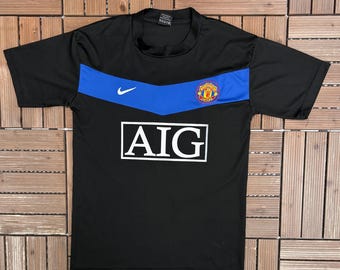 May include: Black short-sleeved jersey with a blue horizontal stripe across the chest. The jersey features the Nike logo, a team crest, and the AIG logo in white. The jersey is likely for sports or athletic wear.