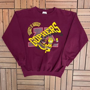 Könnte beinhalten: Ein weinrotes Sweatshirt mit einer Grafik des Maskottchens der University of Minnesota, Goldy Gopher, in Gelb und Schwarz. Das Wort "Gophers" steht in großen gelben Buchstaben über dem Maskottchen. Die Worte "University of Minnesota" sind über dem Wort "Gophers" angeordnet.