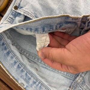May include: Light blue denim jeans with a faded wash. The jeans have a classic five-pocket design and a button and zipper closure. A tag inside the jeans reads "501 S" and the size "34 x 32".