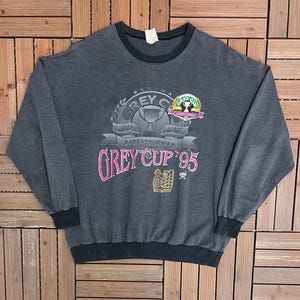 May include: A vintage grey sweatshirt with black stripes and a crew neck. The front features a graphic for the 1995 Grey Cup in Regina, Saskatchewan, with the words "GREY CUP '95" in pink.