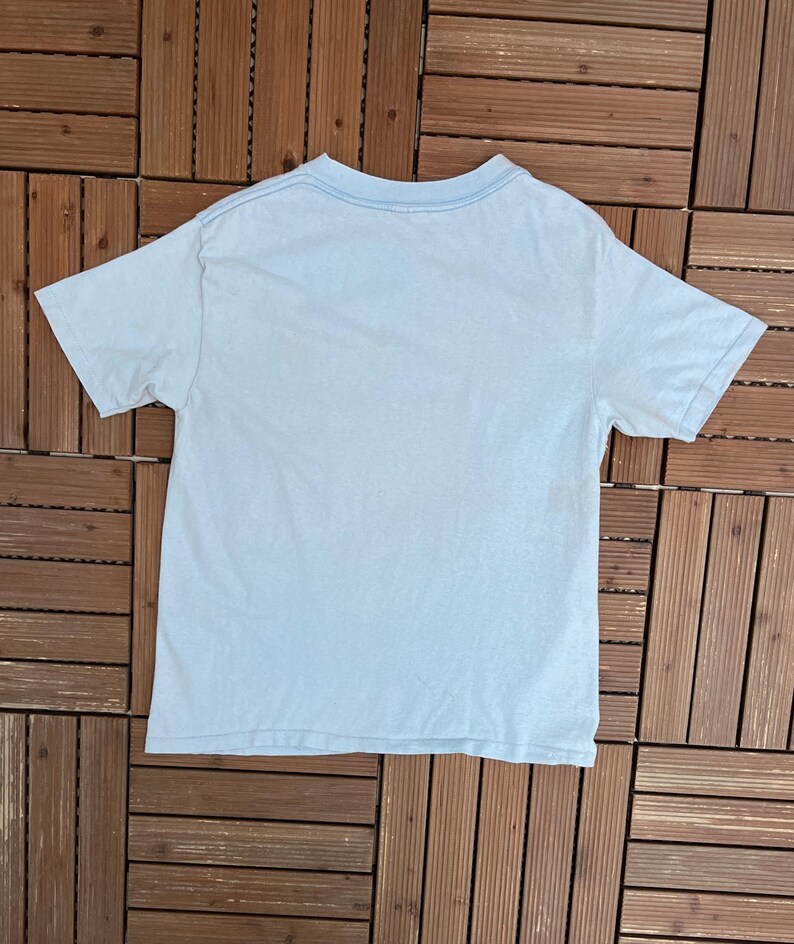 May include: A light blue short-sleeved t-shirt, viewed from the back, with a crew neck. The shirt is made of a soft material and is laid flat on a wooden surface. The shirt's color is a pale, faded blue.