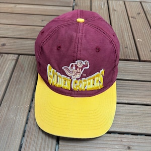 Puede incluir: Una gorra de béisbol granate con visera amarilla y un botón amarillo en la parte superior. El frente presenta un gráfico bordado de una mascota gopher sobre el texto amarillo "GOLDEN GOPHERS" con un pequeño símbolo de marca registrada.
