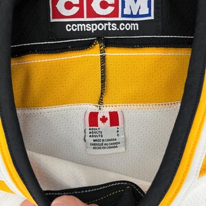 May include: Close-up of a hockey jersey collar with a black, yellow, and white design. The CCM logo is visible, along with the text "ccmsports.com". A tag indicates the jersey was made in Canada.