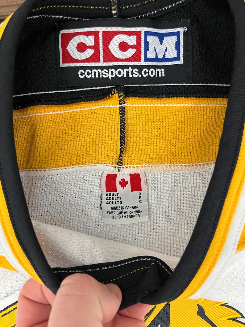 May include: Close-up of a hockey jersey collar with a black, yellow, and white design. The CCM logo is visible, along with the text "ccmsports.com". A tag indicates the jersey was made in Canada.