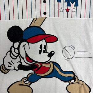 May include: A white baseball-style shirt with thin red and blue pinstripes, featuring three black buttons at the collar. The chest displays the letters MM above three stars. A graphic of Mickey Mouse in a baseball uniform swings a bat at a ball.