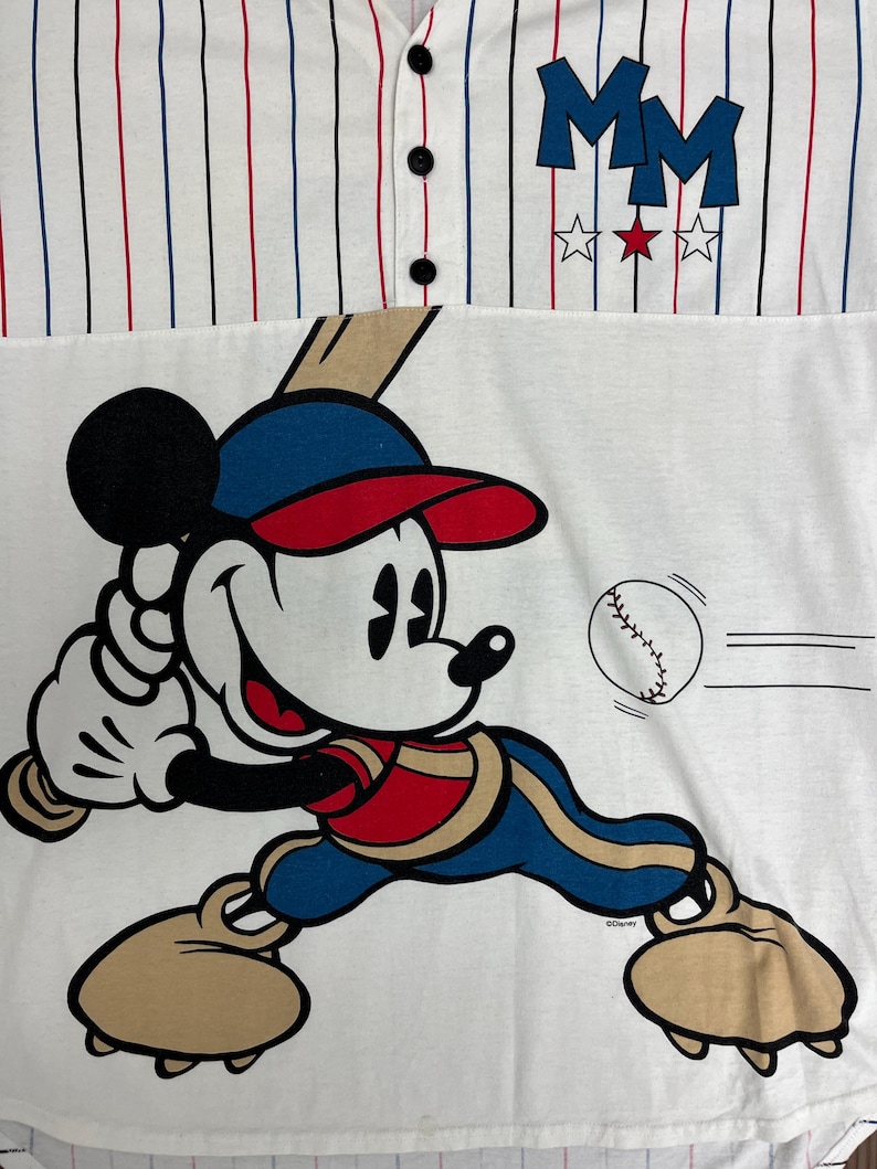 May include: A white baseball-style shirt with thin red and blue pinstripes, featuring three black buttons at the collar. The chest displays the letters MM above three stars. A graphic of Mickey Mouse in a baseball uniform swings a bat at a ball.