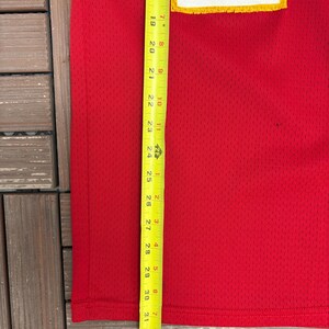 May include: A red mesh jersey with a white and yellow design. A yellow and black measuring tape is placed vertically along the jersey, showing measurements in inches. The jersey is on a wooden surface.