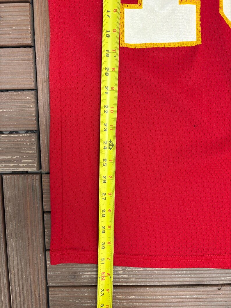 May include: A red mesh jersey with a white and yellow design. A yellow and black measuring tape is placed vertically along the jersey, showing measurements in inches. The jersey is on a wooden surface.