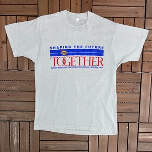 Könnte beinhalten: Hellgraues T-Shirt mit dem Text "SHAPING THE FUTURE" über "TOGETHER" in Blau und Rot. Das Shirt enthält auch "SUNOCO MARKETERS CONFERENCE NEW ORLEANS, LOUISIANA 1989". Das T-Shirt liegt flach auf einer Holzoberfläche.
