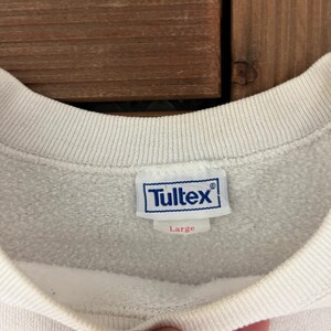 K&ouml;nnte beinhalten: Nahaufnahme eines wei&szlig;en Sweatshirt-Kragens mit einem blau-wei&szlig;en Tultex-Etikett. Das Etikett zeigt die Gr&ouml;&szlig;e Large an. Das Sweatshirt befindet sich auf einer Holzoberfl&auml;che.