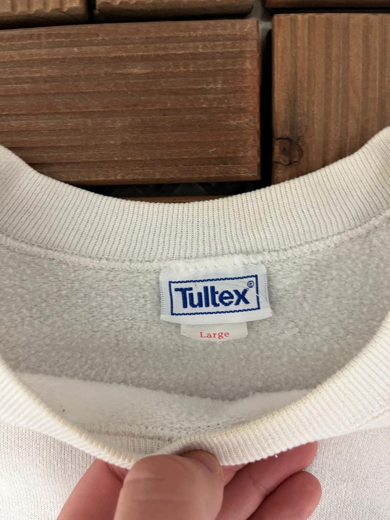 K&ouml;nnte beinhalten: Nahaufnahme eines wei&szlig;en Sweatshirt-Kragens mit einem blau-wei&szlig;en Tultex-Etikett. Das Etikett zeigt die Gr&ouml;&szlig;e Large an. Das Sweatshirt befindet sich auf einer Holzoberfl&auml;che.