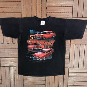May include: Black t-shirt featuring a graphic of red classic Chevrolet cars. The design includes the text "TRI 5 CHEVROLET" and "Classic Iron". The shirt has a crew neck and short sleeves.