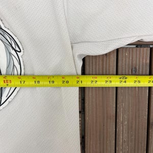 May include: A white jersey with a black bottom hem and a team logo on the upper left. A yellow measuring tape is laid across the jersey, showing measurements in inches. The jersey has a perforated texture.