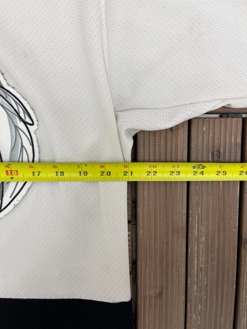 May include: A white jersey with a black bottom hem and a team logo on the upper left. A yellow measuring tape is laid across the jersey, showing measurements in inches. The jersey has a perforated texture.