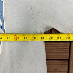 May include: A light blue t-shirt with a white and blue graphic. A yellow measuring tape shows the shirt's width at 16 inches. The shirt is on a wooden surface.