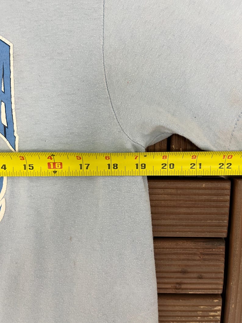 May include: A light blue t-shirt with a white and blue graphic. A yellow measuring tape shows the shirt's width at 16 inches. The shirt is on a wooden surface.