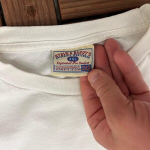 May include: A white t-shirt with a label showing "STEVE & BARRY'S UNIVERSITY SPORTSWEAR XXL Engineered For Comfort MAXIMUM DURABILITY AUTHENTIC STYLING S&B". The label has a blue and red design.