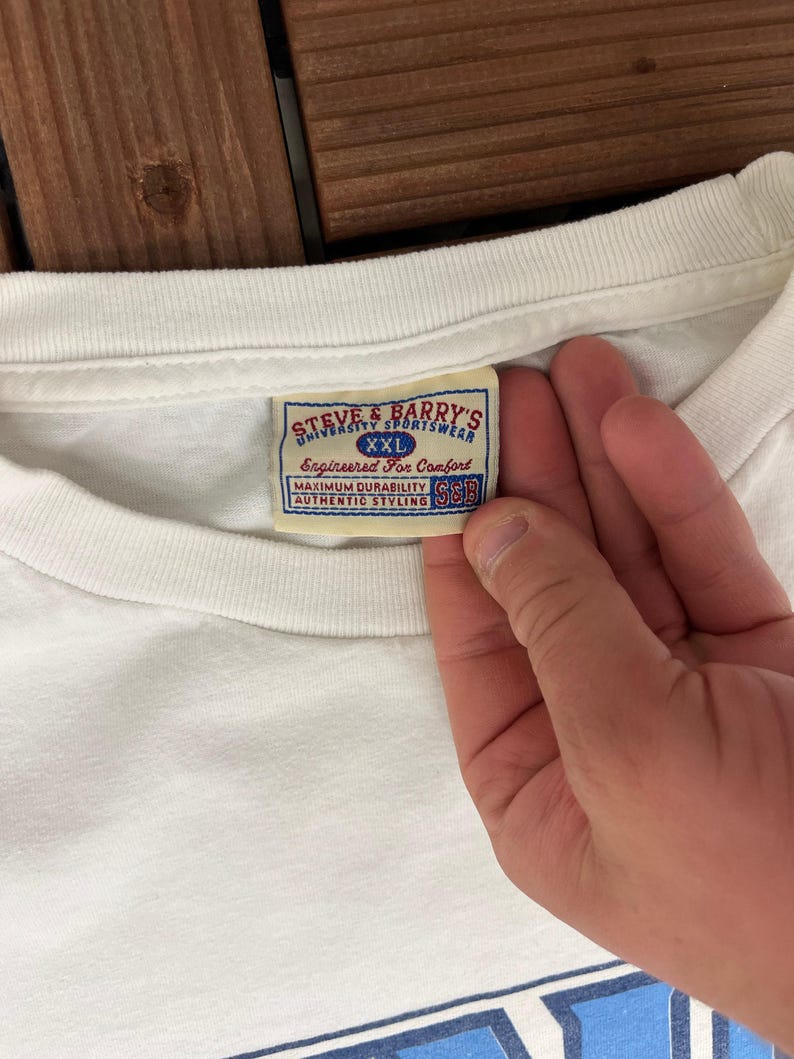 May include: A white t-shirt with a label showing "STEVE & BARRY'S UNIVERSITY SPORTSWEAR XXL Engineered For Comfort MAXIMUM DURABILITY AUTHENTIC STYLING S&B". The label has a blue and red design.