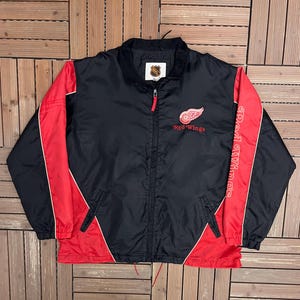 May include: Black and red Red Wings jacket with a full zip. The jacket features the Red Wings logo on the chest and the team name on the right sleeve. The NHL logo is on the collar.