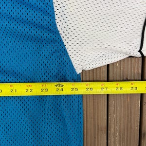 May include: A close-up of a blue, white, and black mesh jersey. The jersey is measured with a yellow tape measure, showing a length of approximately 9 inches. The jersey has a breathable, perforated design.