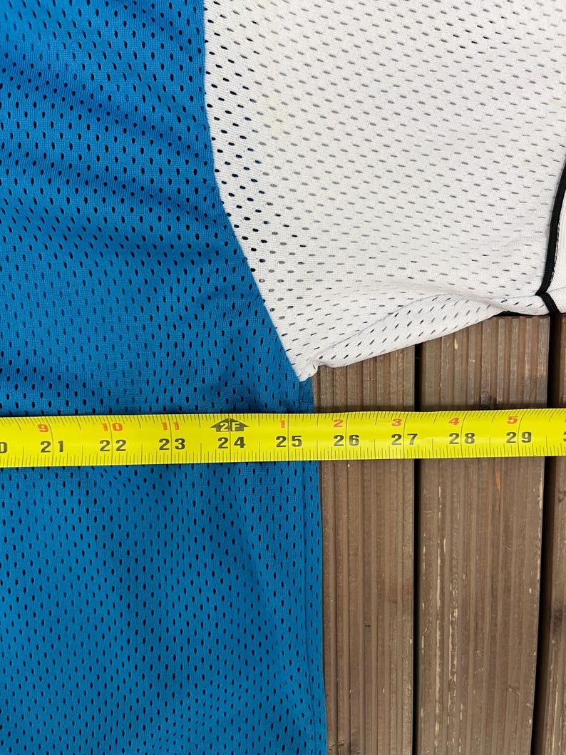 May include: A close-up of a blue, white, and black mesh jersey. The jersey is measured with a yellow tape measure, showing a length of approximately 9 inches. The jersey has a breathable, perforated design.
