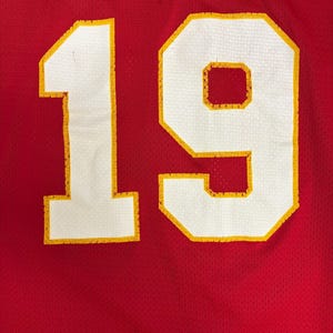 May include: Close-up of a red jersey featuring the number 19 in white with yellow edging. The jersey has a mesh texture. The number is large and centrally positioned, dominating the image.
