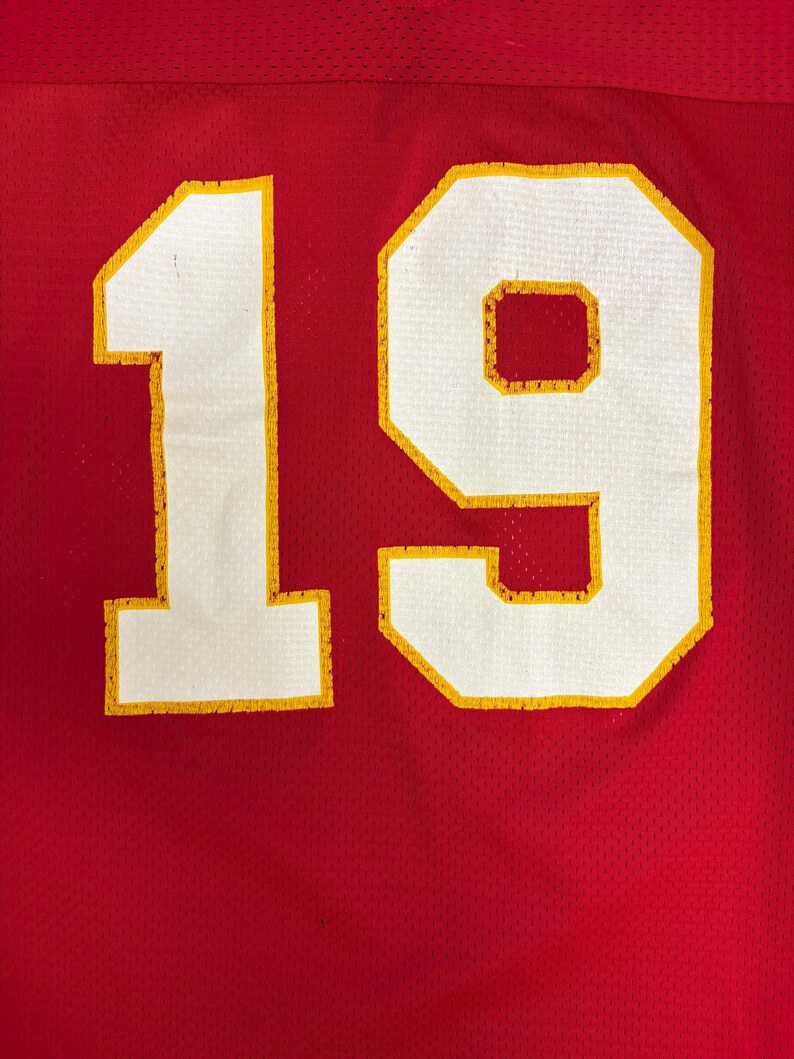 May include: Close-up of a red jersey featuring the number 19 in white with yellow edging. The jersey has a mesh texture. The number is large and centrally positioned, dominating the image.