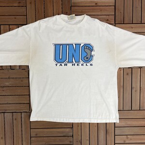 May include: A white long-sleeved t-shirt featuring the text "UNC TAR HEELS" in blue. The shirt has a crew neck and long sleeves. The graphic includes the letters "UNC" and a footprint.