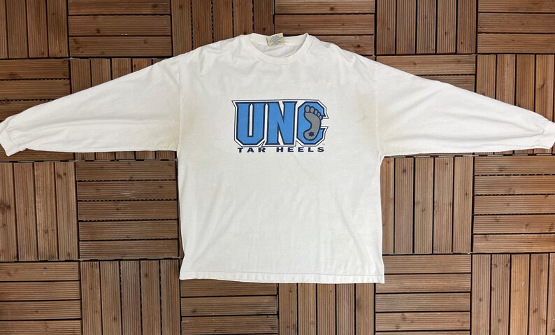 May include: A white long-sleeved t-shirt featuring the text "UNC TAR HEELS" in blue. The shirt has a crew neck and long sleeves. The graphic includes the letters "UNC" and a footprint.