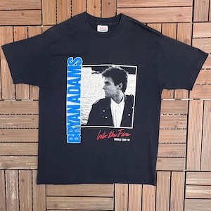 Könnte beinhalten: Schwarzes T-Shirt mit einem Porträt von Bryan Adams und dem Text "BRYAN ADAMS" in Blau. Die Grafik zeigt "Into the Fire World Tour '87". Das T-Shirt besteht aus einem weichen Material und ist ein Vintage-Artikel.
