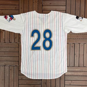May include: A white baseball jersey with thin red and blue pinstripes, featuring the number 28 in large blue digits with gold outlines on the back. The sleeves display small graphic illustrations of a cartoon character wearing a baseball cap.