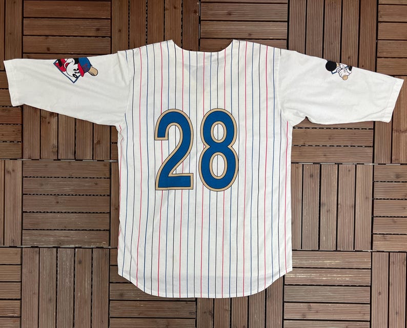 May include: A white baseball jersey with thin red and blue pinstripes, featuring the number 28 in large blue digits with gold outlines on the back. The sleeves display small graphic illustrations of a cartoon character wearing a baseball cap.