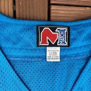 May include: Close-up of a light blue mesh jersey with a black and red logo. The jersey has a V-neck design and a tag that reads "100% Polyester, Size XXL, Made in Korea."