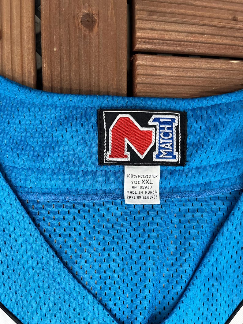 May include: Close-up of a light blue mesh jersey with a black and red logo. The jersey has a V-neck design and a tag that reads "100% Polyester, Size XXL, Made in Korea."