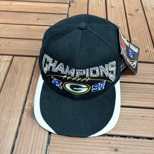 May include: A black baseball cap featuring the word CHAMPIONS embroidered in white and silver thread. Below the text is a Green Bay Packers logo with the number 91 in blue and white. The cap has a white brim edge and an attached product tag.