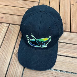 Può includere: Un cappellino da baseball nero con visiera curva. Il cappellino presenta un colorato disegno ricamato di una manta in tonalità blu, verde e giallo. Realizzato in tessuto resistente, ha una vestibilità strutturata.