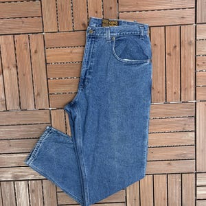 May include: Blue denim jeans with a classic five-pocket design. The jeans have a button and zip closure, belt loops, and a straight leg cut. The denim appears to be a medium wash, with subtle fading. The jeans are laid flat on a wooden surface.