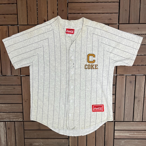 May include: Grey baseball jersey with black pinstripes, featuring a "Coke" logo and a "Coca-Cola" label. The jersey has short sleeves and a button-up front. The jersey is made of a soft, comfortable material.