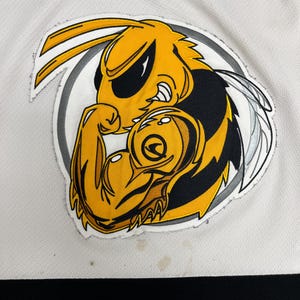 May include: A white jersey with a yellow, black, and white cartoon hornet logo flexing its arm. The jersey has black and yellow stripes at the bottom and a yellow and black V-neck collar. The hornet logo is inside a gray circle.