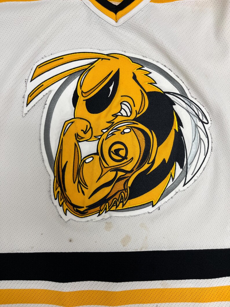 May include: A white jersey with a yellow, black, and white cartoon hornet logo flexing its arm. The jersey has black and yellow stripes at the bottom and a yellow and black V-neck collar. The hornet logo is inside a gray circle.