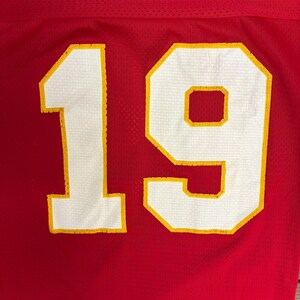 May include: Close-up of a red jersey with the number 19. The number is white with a yellow outline. The jersey has a mesh texture.