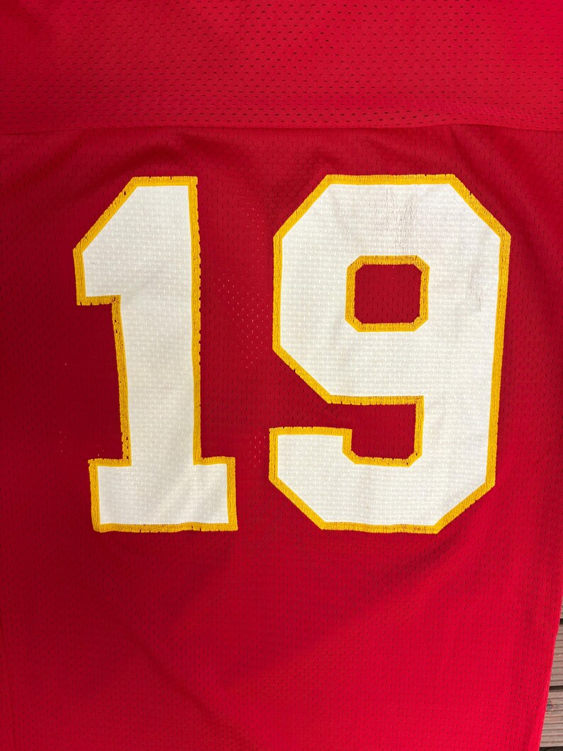 May include: Close-up of a red jersey with the number 19. The number is white with a yellow outline. The jersey has a mesh texture.