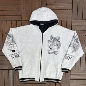 Peut inclure: Un sweat à capuche zippé gris clair avec une doublure intérieure noire. Le sweat-shirt présente un motif de tête de loup détaillé sur le devant et les manches, avec le texte "Canadian Wilderness WHITE WOLF". Bordure rayée noire et blanche.