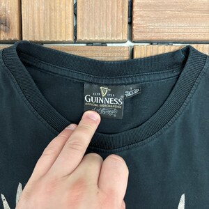 May include: Black t-shirt with a crew neck collar. The shirt features a Guinness Official Merchandise label. The label includes the Guinness logo and the text "ESTD 1759". The shirt is made of a soft material.