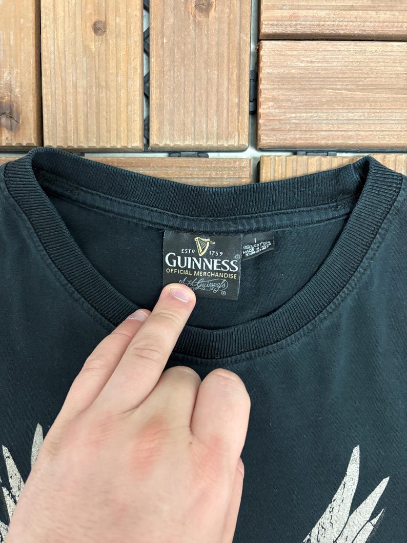 May include: Black t-shirt with a crew neck collar. The shirt features a Guinness Official Merchandise label. The label includes the Guinness logo and the text "ESTD 1759". The shirt is made of a soft material.