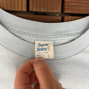 May include: Light blue t-shirt with a Super Shirts tag. The tag reads "100% Cotton, Made in the U.S.A." and has a size "L". The shirt is made of a soft material.