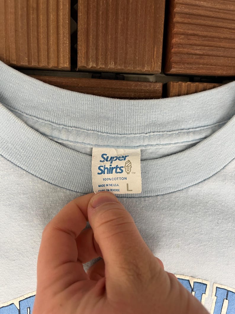 May include: Light blue t-shirt with a Super Shirts tag. The tag reads "100% Cotton, Made in the U.S.A." and has a size "L". The shirt is made of a soft material.
