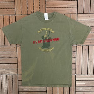 Puede incluir: Camiseta verde oliva con un diseño de la película Monty Python y el Santo Grial. El diseño incluye un caballero y el texto "IT'S JUST A FLESH WOUND" en rojo, con "Monty Python" y "The Holy Grail" en dorado.