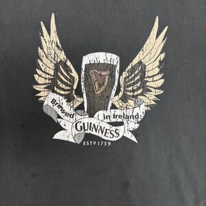 May include: Dark gray t-shirt featuring a Guinness logo with a pint of stout, a harp, and wings. The text reads "Brewed in Ireland" and "Guinness ESTD 1759" in a distressed, vintage style.