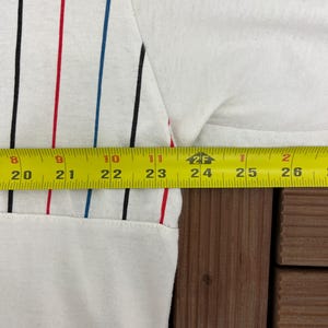 May include: A white shirt featuring vertical pinstripes in black, red, and blue. A yellow measuring tape is positioned across the fabric, showing a width measurement extending from 20 inches to 26 inches across the garment's midsection.